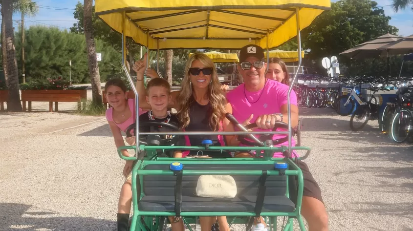Family of five seated on a four‐wheeled surrey bike with a yellow canopy, parked on a gravel lot lined with palm trees and rental bicycles in the background.