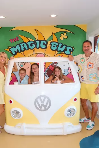 Family of five posing around a white and yellow VW bus photo‐op inside “The Magic Bus” ice‐cream shop, with colorful mural behind them.