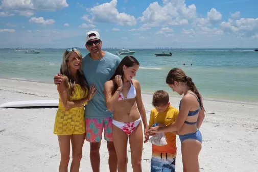 Family of five standing barefoot on a sunny beach in colorful swimwear, with boats on turquoise water and a clear sky behind them.