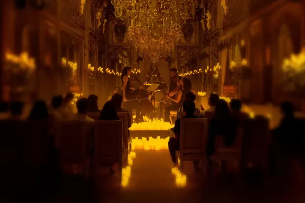 A candlelit classical music concert in an ornate hall with chandeliers and gilded mirrors. A string quartet, dressed in black, performs in the center, surrounded by glowing candles. The audience sits in rows of chairs, attentively watching the musicians. The warm golden lighting enhances the elegant and intimate atmosphere.